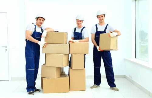 Three male movers dressed in blue overalls, white t-shirts, and white safety helmets are standing inside a bright, modern room with white walls and a large window allowing natural light to fill the space. They are holding and stacking several cardboard boxes of varying sizes, some of which appear to be sealed with packing tape. The boxes have a brown, corrugated texture typical of cardboard used for removals, and they are positioned in a way that suggests the team is in the process of packing or unpacking. The first mover on the left is holding a box with both hands, smiling gently at the camera, while the second in the middle is resting a box on top of a stack of larger boxes, with a calm, focused expression. The third on the right is holding a box against his stomach, also smiling. The floor is a clean, white tiled surface, and the environment appears to be part of a residential property, possibly a living room or hallway, with minimal visible furnishings. The scene conveys professional house removals activity typical for a property in Islington, London, aligned with a local removals service such as Islington Man with Van.