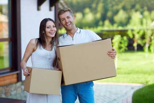 A smiling couple, a woman and a man, standing outside a house in a suburban area with a well-maintained lawn and green trees in the background, each holding a medium-sized cardboard box. The woman has long dark hair and is wearing a sleeveless white dress, while the man has short blonde hair, a beard, and is dressed in a white shirt and blue trousers. The scene appears to be set during daytime with natural light highlighting their faces and the surrounding outdoor environment, indicating a house move or delivery process in an area consistent with Islington or nearby London postcode regions. The house's exterior visible in the background features a brick wall and a window, with part of a driveway or pathway leading up to the home, suggesting a residential setting suitable for a house removals service by Islington Man with Van.