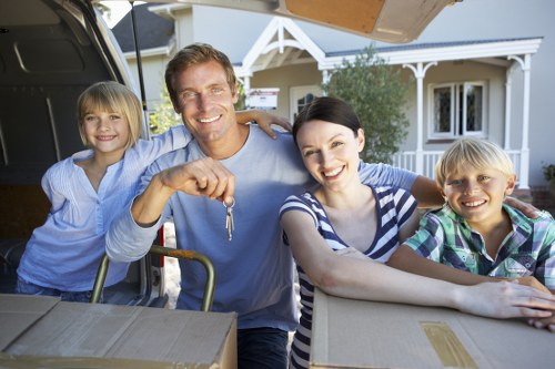 A family of four preparing to load boxes into a black removals van parked in front of a residential house with a white exterior and pitched roof. The father, a smiling man with short brown hair wearing a light blue shirt, is holding a set of keys and standing behind the open van trunk. To his left is a young girl with blonde hair in a ponytail, dressed in a light blue dress, leaning on the van and smiling. On the right, a woman with short black hair, wearing a striped shirt, is leaning on the boxes and smiling at the camera. Beside her, a young boy with blonde hair and a plaid shirt is also smiling. The scene takes place outdoors during daylight, with a driveway or street visible, and is set against a background of a neatly maintained house with a porch, white trim, and greenery. The focus is on the family preparing for a house move, with cardboard boxes stacked nearby, highlighting the house removals theme and conveying a professional, local service context.