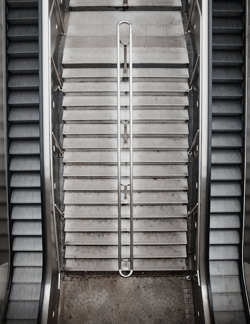 A photograph of a staircase with a central handrail, situated between two escalators in an indoor setting. The stairs are made of light-colored, non-slip material, and the handrail is metallic, extend