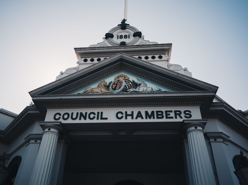 A low-angle view of the facade of a historic council chambers building, featuring classical architectural elements including tall white columns supporting a pediment with decorative relief artwork, a